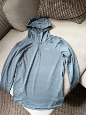 Brooks löparhoodie / träningshoodie - Ljus blågrå träningshoodie från Brooks i storlek S. Teknisk och lätt modell med halvzip, huva, tumhål i ärmsluten och strukturerat material på insidan för bättre ventilation och komfort.  Mycket fint begagnat skick utan tydliga skador eller större slitage. Perfekt för löpning, gym eller annan träning.