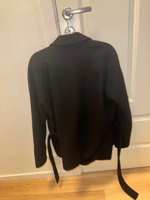 Svart kappa  - Stilren svart kappa med klassisk krage och knytskärp i midjan. Kappan har slät yta och är fodrad på insidan, vilket gör den perfekt för kyligare dagar. Passar till många olika outfits och ger en snygg siluett.