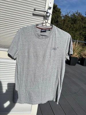 Grå t-shirt från Superdry Athletics - Snygg gråmelerad t-shirt från Superdry Athletics med diskret logga på bröstet. Klassisk rund halsringning och korta ärmar. Tillverkad i mjuk bomull som känns skön mot huden. Perfekt till jeans eller shorts.