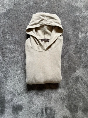 Oscar Jacobson hoodie - En stilren beige hoodie med huva och långa ärmar från Oscar Jacobson. Tillverkad i ett mjukt, stickat material som känns skönt mot huden. Perfekt för dig som gillar en clean och enkel look. Passar till det mesta och är lätt att matcha med andra plagg.