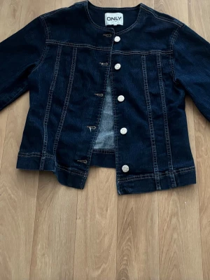 Mörkblå klassisk jeansjacka Gina Tricot  - Snygg mörkblå jeansjacka med raka linjer och vita knappar framtill. Jackan har långa ärmar, markerade sömmar och en enkel, tidlös design. Perfekt för dig som gillar en clean och stilren look. 
