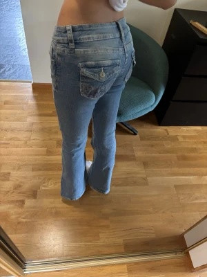 Ljusblå bootcut jeans med fickdetaljer - Säljer ett par ljusblå bootcut jeans med snygga fickdetaljer och knappar bak. Tre hål där, lätt tvättade. Finns inga skador och har inte använts mycket. 