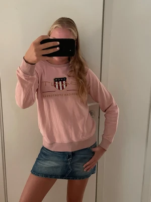 Ljusrosa sweatshirt med tryck - Säljer en ljusrosa sweatshirt med tryck och emblem framtill. Tröjan har rund halsringning, ribbade muddar vid ärmslut och nederkant samt en avslappnad passform. Perfekt för en casual look och enkel att matcha med jeans eller kjol.