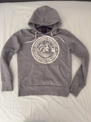 Superdry hoodie - Snygg grå hoodie från Superdry med stort vitt tryck på bröstet där det står 'Superdry Everest Expedition Company' och ett bergmotiv. Hoodien har huva med vita snören och en klassisk magficka. Perfekt för en avslappnad och sportig stil.