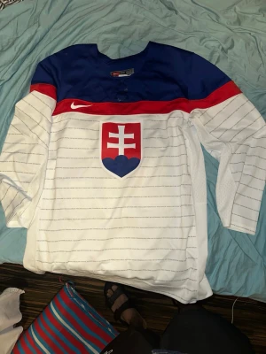Slovakisk hockeytröja Nike vit/blå - Säljer en vit hockeytröja från Nike med Slovakiens landslagsemblem på bröstet, blå axelparti och röd rand över bröstet. Tröjan har blåa detaljer, nummer 83 på ryggen och IIHF-märke. Perfekt för hockeyfans som vill sticka ut på matchen.