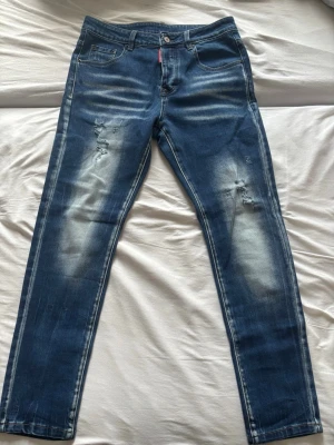 Mörkblåa dsquared 2 jeans - Säljer ett par helt oanvända blå dsquared 2 jeans med slitningar på framsidan och ljusa detaljer. Jeansen har raka ben, klassiska fickor och en knappstängning. Materialet är denim och de har en cool, lite vintage look.