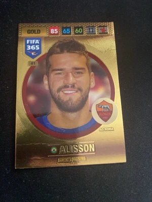 FIFA 365 Gold Alisson spelkort - Unikt FIFA 365 Gold samlarkort med Alisson från AS Roma. Kortet har en guldig bakgrund, färgstarka stats och klubbmärke. Perfekt för dig som gillar fotboll och samlarkort. Kortet är gjort av glansig kartong och har snygg design.