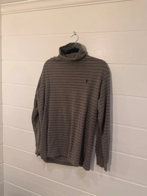 Grå Randig polotröja från Polo Ralph Lauren Storlek L - Mjuk och skön polotröja från Polo Ralph Lauren i grått med tunna svarta ränder. Tröjan har lång ärm, hög krage och en liten broderad logga på bröstet. Perfekt för lager på lager och en avslappnad stil.