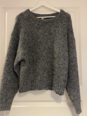 Grå fluffig stickad tröja Nelly - Mysig grå stickad tröja från Nelly med fluffig textur och avslappnad passform. Tröjan har rund halsringning och långa ärmar, perfekt för kyliga dagar. Enkel och stilren design som är lätt att matcha med olika outfits.