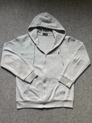 Grå zip hoodie från Polo Ralph Lauren - Snygg grå hoodie med dragkedja från Polo Ralph Lauren. Klassisk design med huva och dragsnören, två fickor framtill och den ikoniska svarta loggan broderad på bröstet. Tillverkad i mjuk bomullsblandning som är perfekt för chill dagar.