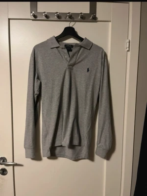 Grå långärmad piké från Polo Ralph Lauren - Säljer en stilren grå långärmad pikétröja från Polo Ralph Lauren med klassisk krage och knappar framtill. Tröjan har det ikoniska blå logobroderiet på bröstet och är gjord i mjuk bomull. Perfekt för en clean och avslappnad look.