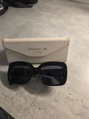 Svarta solglasögon från STOCKH LM Studio - Snygga svarta solglasögon från STOCKH LM Studio med breda, fyrkantiga bågar. Glasen är mörka och bågarna har en stilren, modern look. Perfekta för att höja din outfit och skydda ögonen mot solen.