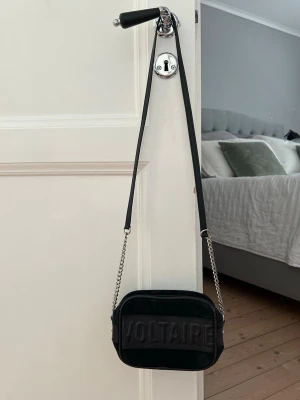 Svart handväska från Zadig & Voltaire - Snygg svart handväska i mocka från Zadig & Voltaire med präglad logga framtill. Väskan har silverfärgade kedjedetaljer på axelremmen och en kompakt, rektangulär form. Perfekt för dig som gillar stilrena accessoarer med en edgy touch.
