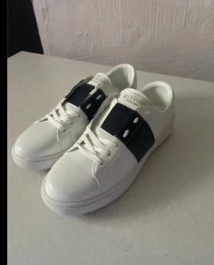 Vita sneakers med svart detalj - Snygga vita sneakers från Valentino med bred blå läderdetalj över snörningen. Klassisk låg modell med rund tå och vita skosnören. Märkeslogga på plösen och stilren design som passar till det mesta.