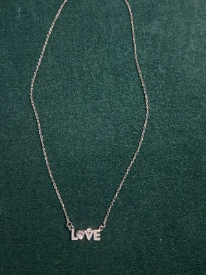Silverhalsband med LOVE-bokstäver - Snyggt halsband i silverfärg med tunnt kedja och bokstäverna LOVE som hänge. Bokstäverna är dekorerade med små gnistrande stenar och ett hjärta istället för o. 