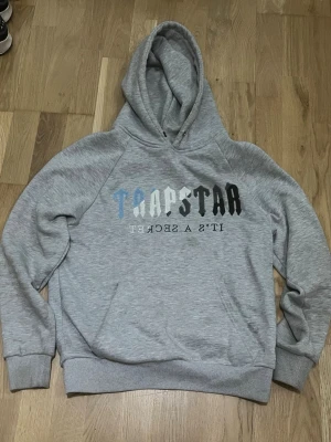 Grå hoodie Trapstar - Snygg grå hoodie med stor huva och Trapstar-tryck i flera färger på bröstet. Texten 'IT'S A SECRET' står under loggan. Hoodien har känguruficka framtill och ribbade muddar vid ärmar och nederkant. Perfekt för en avslappnad streetwear-look.