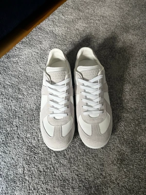 Margiela sneakers  - Snygga sneakers i vitt och ljusgrått med ovandel i en mix av mocka och läder. Klassisk siluett med vita skosnören och diskreta detaljer. Perfekta för en clean och stilren look.