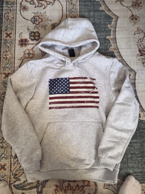 Grå hoodie med amerikansk flagga - Snygg ljusgrå hoodie med stor amerikansk flagga tryckt på bröstet. Hoodien har klassisk känguruficka, ribbade muddar och justerbar huva med dragsko. Perfekt för dig som gillar streetwear och vill sticka ut lite extra. Storlek S personen på bilden är 178