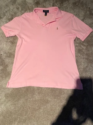 Rosa pikétröja från Polo Ralph Lauren - Snygg rosa pikétröja från Polo Ralph Lauren med klassisk krage och korta ärmar. Tröjan har den ikoniska lilla broderade loggan på bröstet och är tillverkad i mjuk bomull. Perfekt för en avslappnad men stilren look.