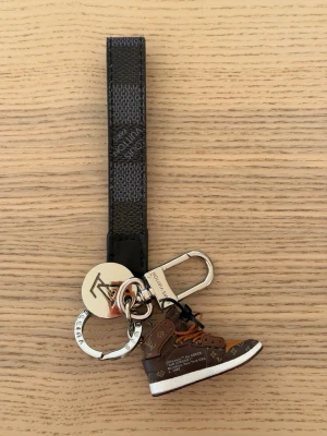  Vuitton nyckelring sneaker - Snygg nyckelring från  Vuitton med en miniatyr av en brun sneaker, detaljer i orange och vitt samt LV-logga. Metallringar och karbinhake i silverfärg, samt svart band med präglad logotyp. Perfekt accessoar för att piffa till nyckelknippan.