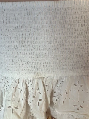 Vit kort kjol med volanger och broderi - Supersöt vit kort kjol med bred smock i midjan och dubbla volanglager. Kjolen har broderade hålmönster och ger en romantisk vibe. Perfekt att styla med en stickad tröja eller crop top för en fräsch look.