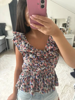 Blommig volangtopp med v-ringning - Supersöt blommig topp med volanger och v-ringning. Toppen har ett färgglatt mönster i rosa, blått och lila, samt en smickrande peplumform. Perfekt att styla med jeansshorts för en fräsch look. Passar som både m och s💕💕
