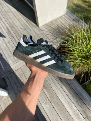 Adidas Spezial mörkgröna sneakers - Säljer ett par Adidas Spezial sneakers i mörkgrönt mocka med vita detaljer och klassiska tre ränder på sidan. Skorna har svart snörning, vit hälflik och gummisula. Guldtexten 'Spezial' syns på sidan. Perfekta för dig som gillar retrostil.