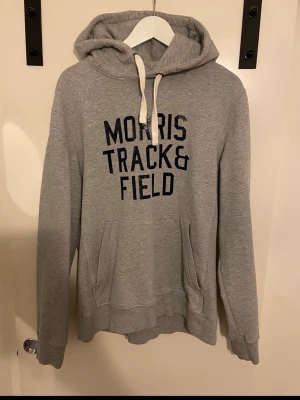 Grå hoodie Morris Track & Field - Snygg grå hoodie från Morris med trycket 'Morris Track & Field' i mörkblått på bröstet. Hoodien har huva med vita snören och en stor magficka framtill. Tillverkad i mjukt bomullsmaterial, perfekt för en avslappnad stil. Storlek L