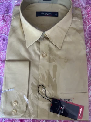 Beige skjorta XL - Klassisk beige skjorta från Uniworth med lång ärm och knappar framtill. Skjortan har en stilren design med enkel bröstficka och normal passform. Perfekt för dig som gillar en clean och tidlös look.