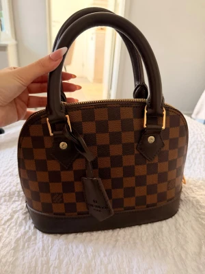 Brun rutig handväska med gulddetaljer - Snygg handväska i brunt rutigt mönster med mörkbruna detaljer och guldfärgade metalldelar. Väskan har två stadiga handtag och dragkedja upptill. Klassisk bågformad modell som ger en lyxig känsla. Perfekt för dig som gillar stilrena accessoarer.