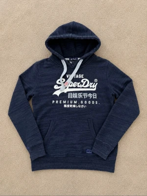 Mörkblå Superdry hoodie med tryck - Snygg mörkblå hoodie från Superdry med vit logga och japanskt tryck på bröstet. Klassisk känguruficka framtill och justerbar huva med vita snören. Tillverkad i mjukt bomullsmaterial som är perfekt för chill dagar. Passar dig som gillar streetwear och enkel stil.