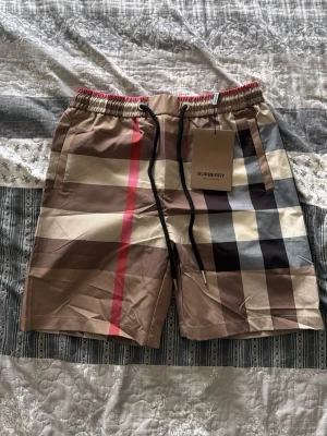 Rutig shorts med snörning från Burberry - Säljer ett par klassiska Burberry shorts med det ikoniska rutiga mönstret i beige, svart, brun och röd. Shortsen har elastisk midja med svart snörning och fickor på sidorna. Perfekta för sommaren eller hemma och ger en lyxig vibe till din outfit.