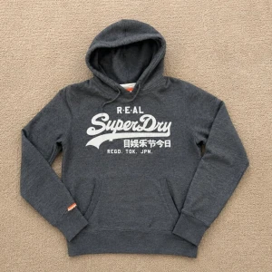 Mörkgrå Superdry hoodie med tryck - Mörkgrå hoodie från Superdry med stor vit logga och japansk text på bröstet. Klassisk huva, magficka och ribbade muddar. Mjuk bomullsblandning och avslappnad passform. Perfekt för en chill och sportig stil.