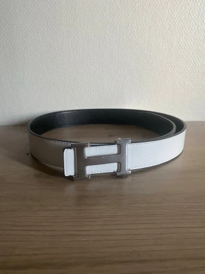 Vändbart svart och vitt bälte med H-spänne - Snyggt vändbart bälte i svart och vitt med ett stort H-format metallspänne. Bältet har en stilren och modern look, perfekt för att lyfta vilken outfit som helst. Tillverkat i skinn med slät yta och silverfärgat spänne.