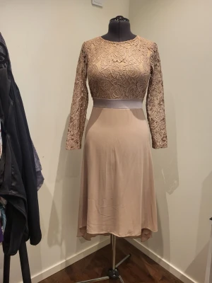 Beige spetsklänning med lång ärm - Elegant beige klänning med spetsliv och långärmade spetsärmar. Kjolen är i mjukt tyg och har en lätt asymmetrisk fåll. Midjan markeras av ett brett satinband med rosett bak. Perfekt för dig som vill ha en romantisk look. Bröst ca 98cm, midja ca 77cm, höft ca 97-100cm, längd från axel ca 105cm framtill, ärm ca 55cm.