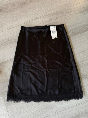 Svart kjol med spetskant mango - Stilren svart kjol i mjukt material med dekorativ spetskant längst ner. Kjolen har en rak passform och är perfekt för dig som gillar en enkel men ändå elegant look.