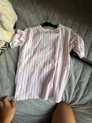 Vit t-shirt med rosa ränder - Säljer en söt vit t-shirt med tunna rosa vertikala ränder och diskreta små rosetter i mönstret. T-shirten har korta puffärmar och rund halsringning. Perfekt för en avslappnad och trendig look till sommaren.