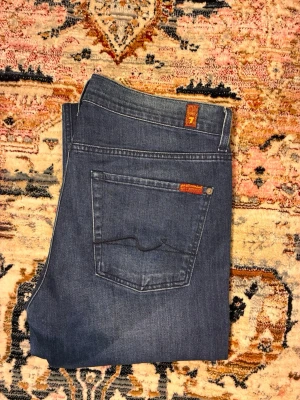 7FAM jeans  - Snygga blå slim fit jeans från 7 For All Mankind med klassisk femficksdesign och subtila slitningar. Jeansen har raka ben och detaljer i orange på bakfickan. Perfekta för en avslappnad och trendig look.