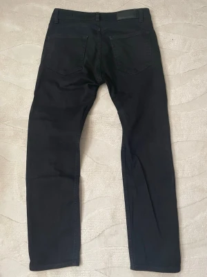 Svarta Dreassman jeans - Säljer ett par svarta jeans med rak passform och klassiska bakfickor. Jeansen har bälteshällor och är tillverkade i ett mjukt denimtyg som sitter snyggt. Perfekta för dig som gillar en clean och stilren look.