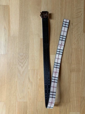 Burberry bälte reversible - Snyggt bälte från Burberrymed rutigt mönster  med beige på ena sidan och svart med diskret mönster på andra sidan. Bältet har ett metallspänne och passar perfekt för att lyfta din outfit.