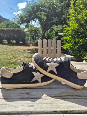 Svarta Golden goose sneakers med stjärndetalj - Snygga svarta golden goose sneakers med vit stjärna på sidan och beige häl. Skorna har vita snören och sula, samt en låg profil. Skorna är gjorda av svart mocka. Perfekta för en avslappnad och trendig stil. Priset kan sänkas vid en snabb affär.