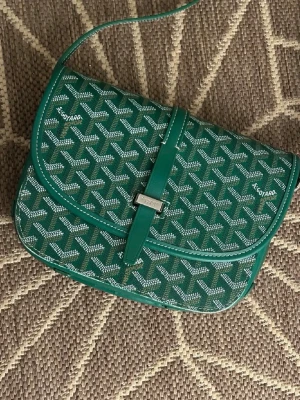 Grön Goyard handväska med mönster - Snygg grön handväska från Goyard med det klassiska vita och beige Goyard-mönstret. Väskan har ett lock med spänne i silverfärgad metall och justerbar axelrem. Tillverkad i canvas med läderdetaljer. Perfekt för dig som vill sticka ut med en ikonisk accessoar.