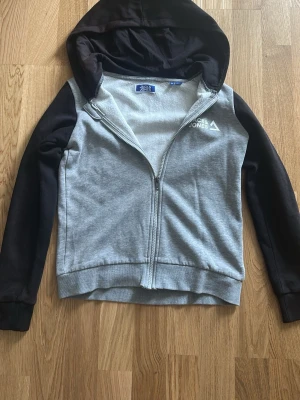 Jack & Jones hoodie - Snygg hoodie från Jack & Jones i grått med svarta ärmar och huva. Dragkedja framtill och mjukt fleecefoder på insidan. Perfekt för chill dagar eller när du vill ha något bekvämt och stilrent.