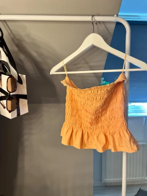 smockad topp  - Säljer en söt persikofärgad topp med smockad överdel . Toppen har en volangkant nedtill som ger en extra touch. Perfekt för sommardagar och enkel att matcha med jeans eller kjol.är bandou top
