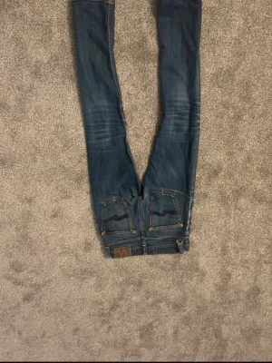Blå smala jeans från Nudie Jeans - Säljer ett par blå smala jeans från Nudie Jeans med klassiska bakfickor och snygga slitningar. Jeansen har en normal midja och är tillverkade i denim med stretch för extra komfort. Perfekta för dig som gillar en avslappnad men ändå stilren look.