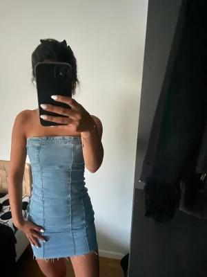 Axelbandslös jeansklänning - Trendig axelbandslös jeansklänning i kort modell med rå kant nertill. Klänningen har en tight passform och markerade sömmar framtill för en cool look. Perfekt för dig som vill ha en enkel men snygg outfit.