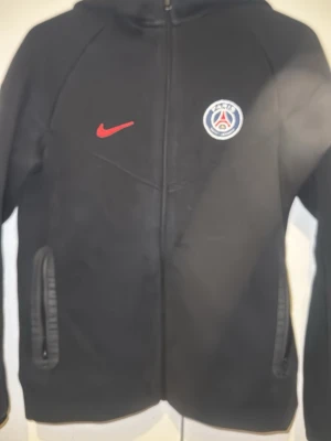 Svart PSG hoodie med dragkedja - Svart hoodie från Nike med PSG-logga och röd swoosh på bröstet. På ryggen står det 'PARIS' i rött. Hoodien har dragkedja framtill, två fickor och huva. Perfekt för dig som gillar fotboll och vill ha en sportig stil.