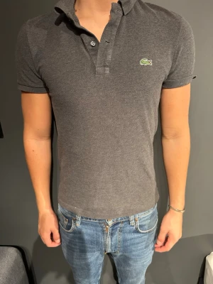 Mörkgrå pikétröja från Lacoste - Snygg mörkgrå pikétröja från Lacoste med klassisk krage och två knappar framtill. Tröjan har korta ärmar och den ikoniska gröna krokodilloggan på bröstet. Perfekt till jeans eller shorts för en clean look. Modellen är 175cm och väger 70kg