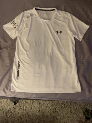 Vit träningströja från Under Armour - Snygg vit t-shirt från Under Armour med reflekterande detaljer och logga på bröstet. Tröjan har rund hals, korta ärmar och tryck på sidan samt ärmen. Perfekt för träning och sportaktiviteter, tillverkad i ett lätt och snabbtorkande material.