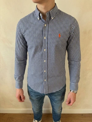 Blårutig skjorta från Polo Ralph Lauren (Slim Fit) - Klassisk blårutig skjorta från Ralph Lauren med button-down krage och lång ärm. Skjortan har vita knappar framtill och en orange broderad logga på bröstet. Tillverkad i mjuk bomull och har en normal passform. Perfekt till jeans eller chinos. Storlek M slim fit. Snabb Leverans!